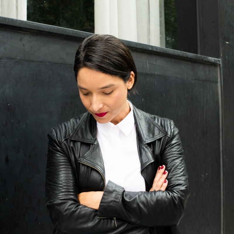 Antonieta wears a black leather jacket and white shirt, stands with arms crossed looking down 